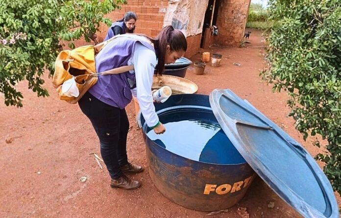 Two people collect water in a rural area, one leaning over a blue water drum with the lid off and pouring from a bottle into the barrel.