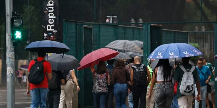 Crowd of pedestrians walking in the rain with colorful umbrellas beside a green construction fence.