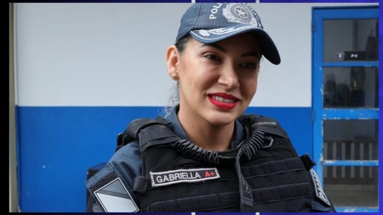 Smiling female police officer in uniform with name patch “GABRIELLA A+,” wearing a cap, standing near a blue wall.