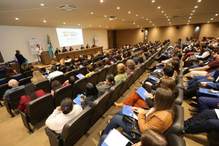 Conference hall with a panel table at the front and a speaker at a podium, addressing a large audience seated in curved rows; a projection screen shows a presentation next to a Brazilian flag.
