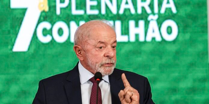 Older man with white beard in a dark suit speaks at a microphone on a stage with a green backdrop and text behind him.