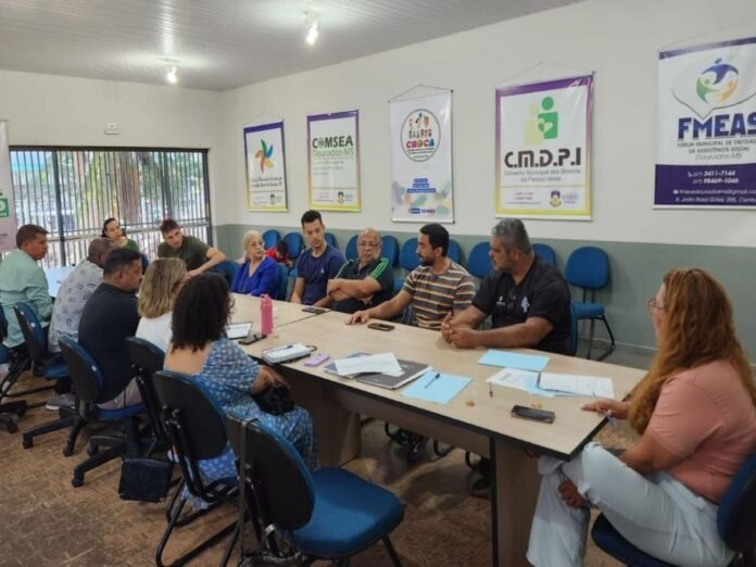 Group of adults seated around a long conference table in a meeting room, discussing documents with posters on the wall behind them.