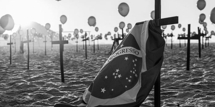 Beach memorial with crosses, balloons, and a Brazil flag draped over a cross in the foreground (black and white).