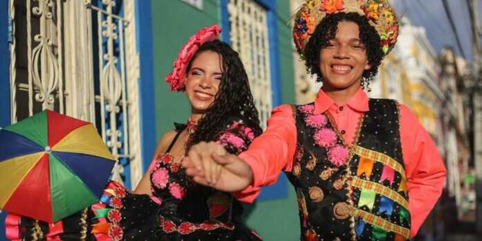 Two smiling women in bright carnival outfits dance hand in hand on a sunny street, a rainbow umbrella nearby.