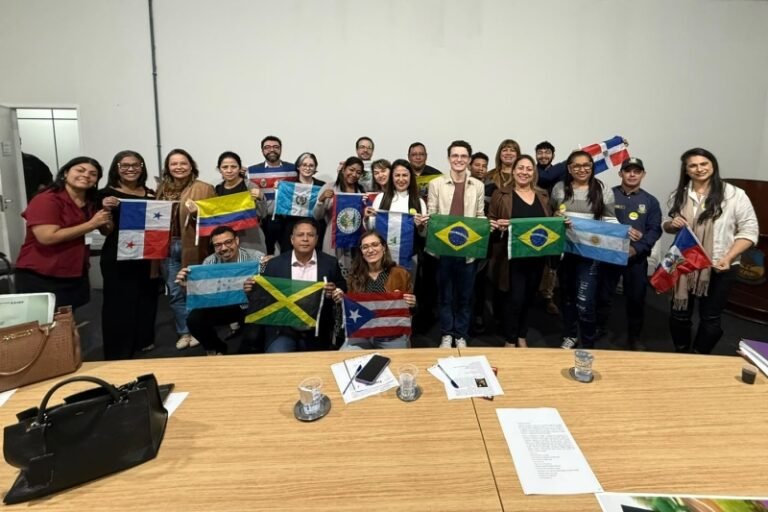 Group of diverse adults in a meeting room posing for a photo while holding multiple national flags (Brazil, Argentina, Jamaica, Panama, Puerto Rico, etc.). It looks like an international or cultural event.