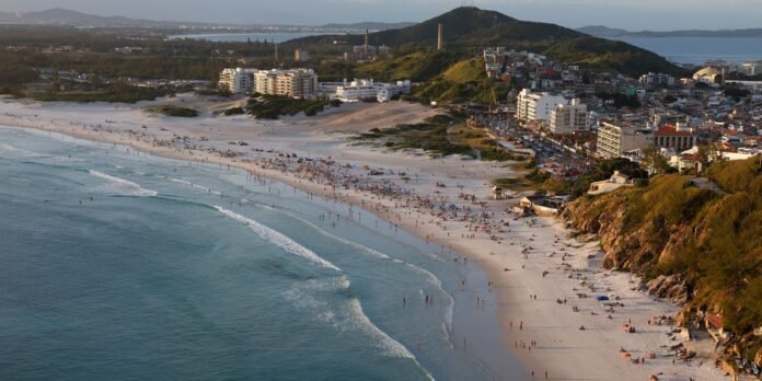 Aerial view of a long sandy beach beside a coastal town with low-rise buildings, hills, and blue ocean waves.