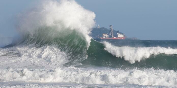 A massive ocean wave curling and crashing with spray, a red ship visible on the horizon in the background.
