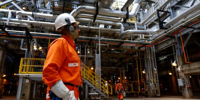 Worker in orange coveralls and a white hard hat stands among a network of metal pipes in a factory.