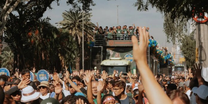 Massive crowd raises hands along a city street as a decorated parade truck passes by with people waving from the top.
