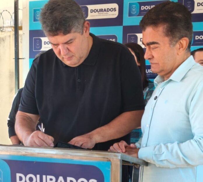 Two men sign a document at a podium during a Dourados Prefeitura event, blue banners behind them.