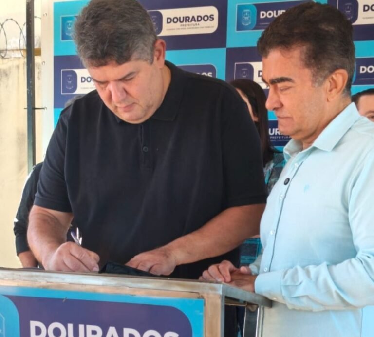 Two men sign a document at a podium during a Dourados Prefeitura event, blue banners behind them.