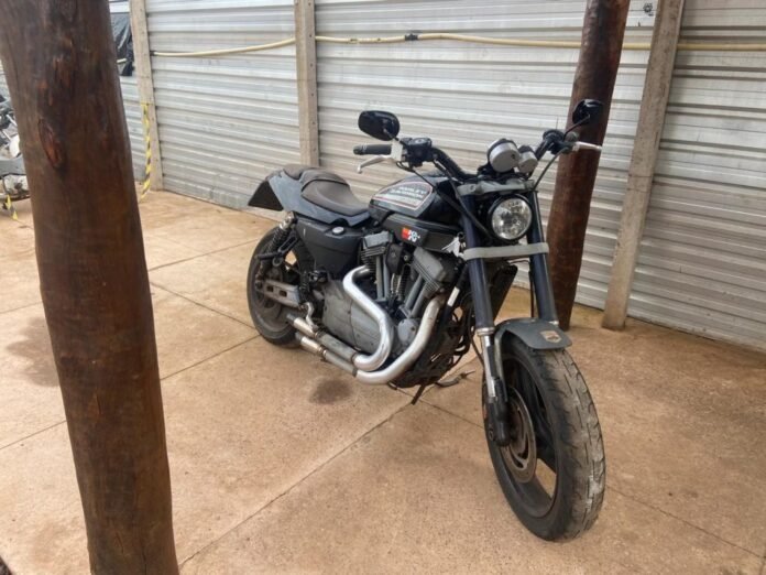 Black Harley-Davidson sport cruiser motorcycle parked on a concrete surface beside rusty poles and a corrugated metal wall/garage backdrop.