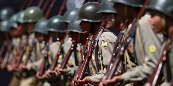 Row of soldiers in khaki uniforms and green helmets kneeling in formation, each holding a rifle with bayonet attached, Brazilian patches visible on sleeves.