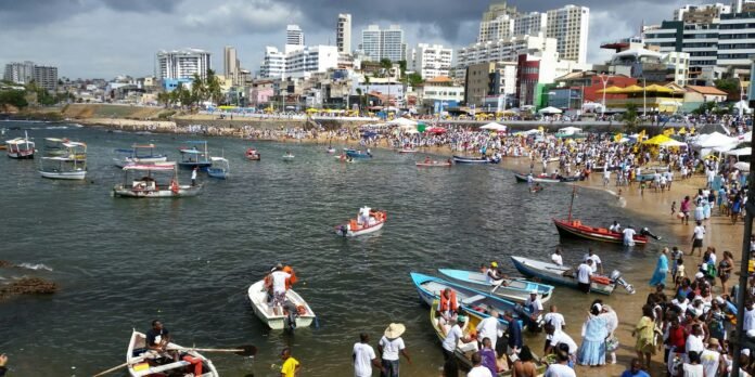 Crowded beachfront festival with small boats in harbor and high-rise buildings in the background.