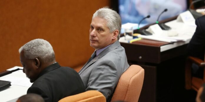 Man in a gray suit sits at a conference table, turning to look at the camera while another man sits beside him and microphones sit on the desk behind them.