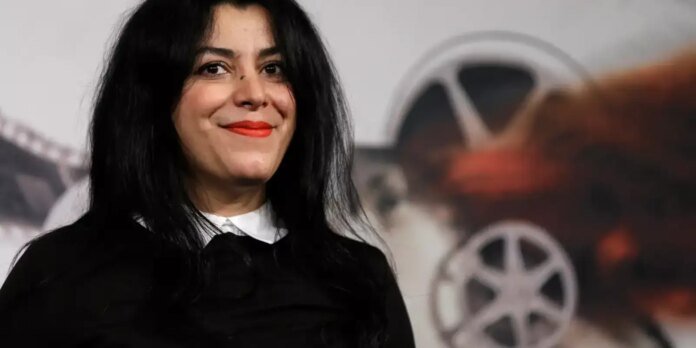 Smiling woman with long black hair, red lipstick, wearing a black top with a white collar, posed in front of a film reel backdrop.
