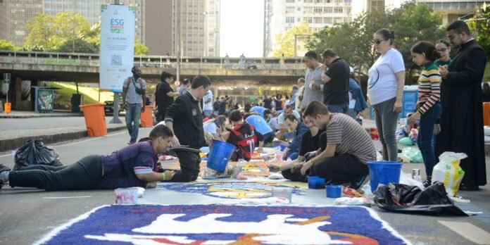 People kneeling and lying along a large painted street carpet as others watch at an urban outdoor art event.