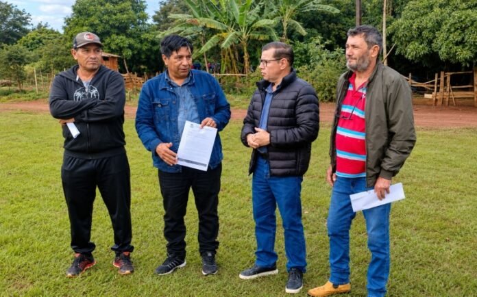Four men stand on a grassy field outdoors, one holding a document while the others listen and talk.