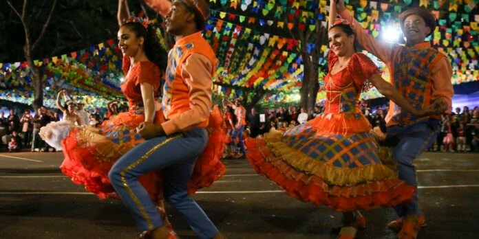 Dancers in bright orange dresses and blue plaid skirts perform a festive street dance under colorful hanging banners.