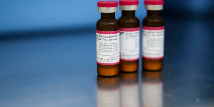 Three small brown vaccine vials with red caps and pink-edged labels sit on a reflective metal surface.
