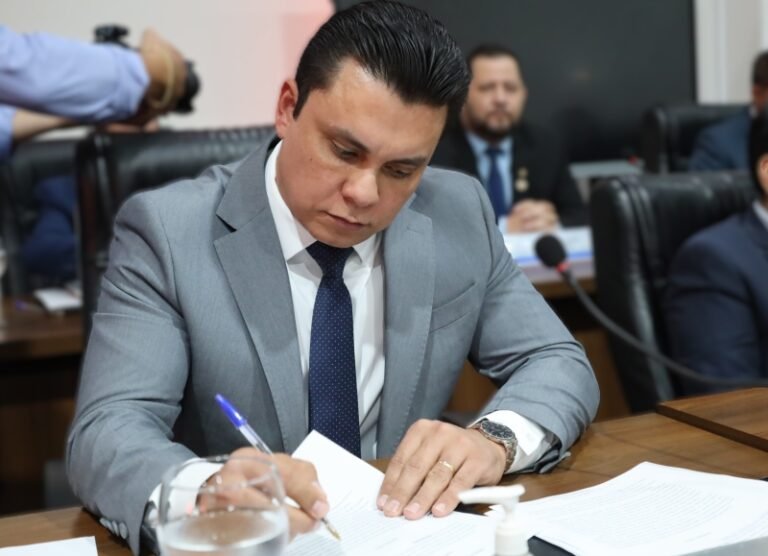 Man in a gray suit signs papers at a conference table, a blue tie visible, with other attendees blurred in the background in a meeting room.