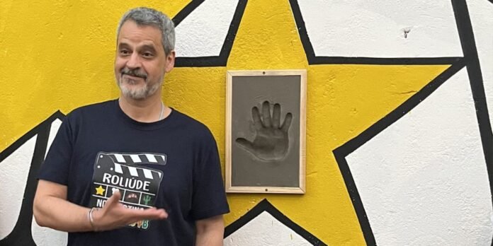 Smiling man in a black T-shirt stands beside a framed handprint impression on a yellow wall with a large star mural.