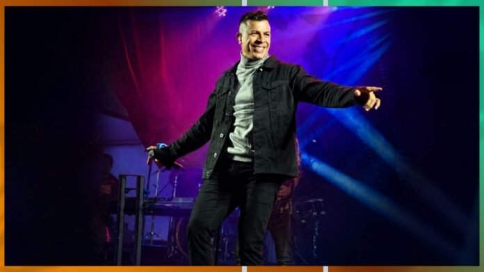 Smiling male singer on stage, wearing a black jacket and gray turtleneck, pointing to the audience as colorful lights shine behind him.