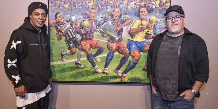 Two men stand on either side of a large framed painting of soccer players celebrating with trophies in a stadium scene.
