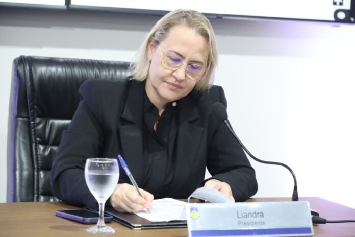 Woman in a black blazer signs papers at a desk, with a microphone and a glass of water nearby.