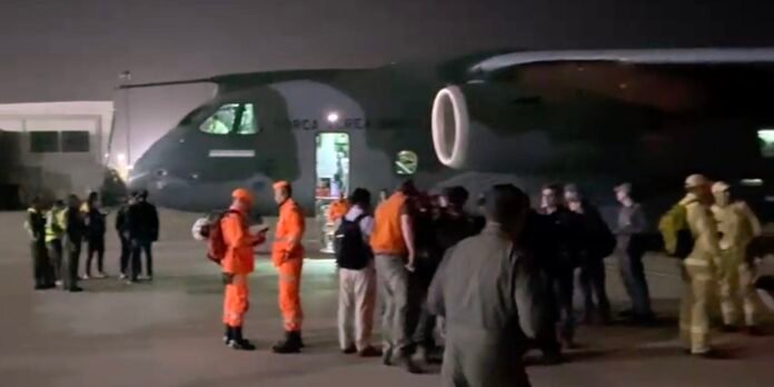 Nighttime at an airstrip with a large dark military transport aircraft and a crowd of people, some in orange safety suits, near its open door.