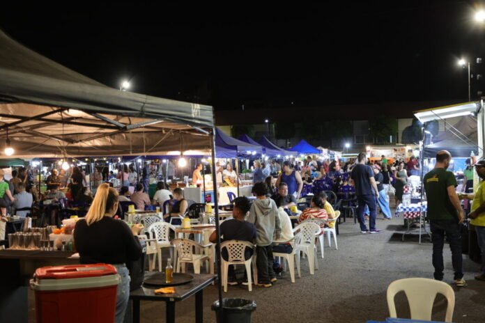 Night market scene with stalls under colorful tents, crowds seated at tables eating by warm lights at night.