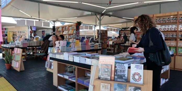 Outdoor book market stall with tables of stacked books under a white canopy; people browse and read.