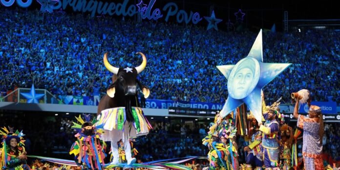 Carnival parade in a stadium: a large black bull costume with colorful fringe, surrounded by dancers in bright outfits.