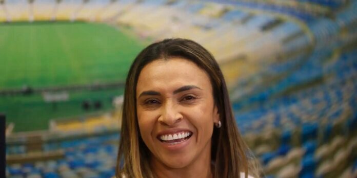 Smiling woman with long brown hair, stadium seating and a green field blurred in the background.