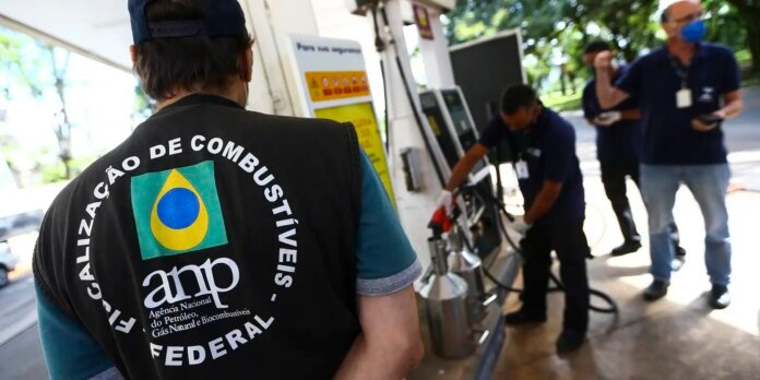 Worker wearing a black vest with Brazilian flag and ANP logo stands at a fuel pump as others refuel a vehicle nearby at an outdoor station.