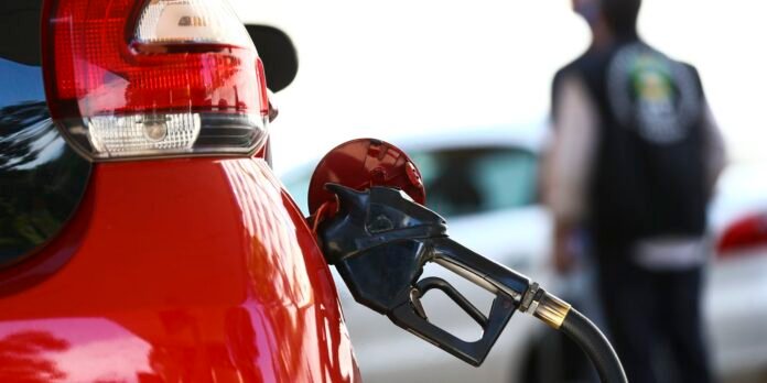 Close-up of a red car being refueled with a black gas pump nozzle inserted into the fuel door, foreground in focus and a blurred person in the background.