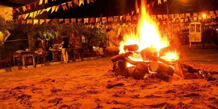 Large bonfire on a sandy beach at night with colorful pennant banners and people gathered in the background.