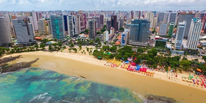 Aerial view of a tropical city shoreline with tall modern buildings along a sandy beach, turquoise water and rows of colorful beach umbrellas.