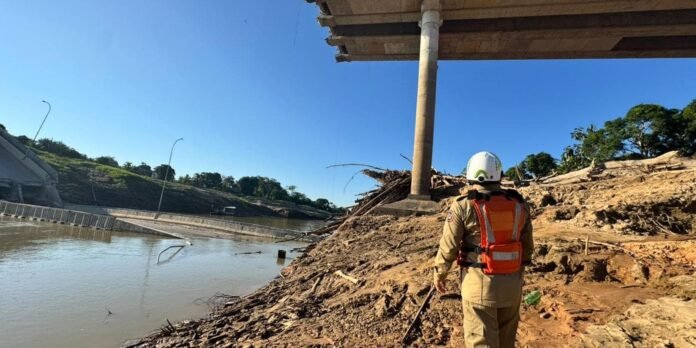 A firefighter in beige uniform and bright orange vest stands on a muddy embankment beside a river under a bridge pillar, surveying a collapsed area with debris.