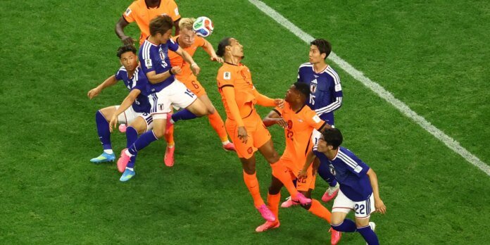 Soccer players from orange and blue teams leap for a header during a match on a green pitch.