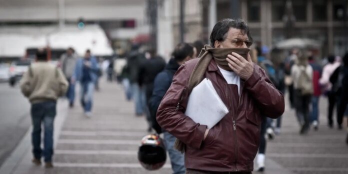 Man in a maroon jacket covers his mouth with a brown scarf while walking on a busy city street, holding papers under his arm.