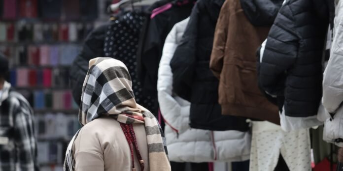 Person wearing a beige coat and a large striped scarf stands near a rack of puffer jackets on a street market.