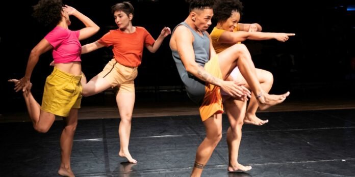 Group of five dancers mid-performance on a dark stage, wearing bright tops and shorts, lifting legs and arms for a dynamic pose to the audience.