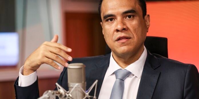 Man in a dark suit and light blue tie speaks into a large studio microphone during a podcast recording.
