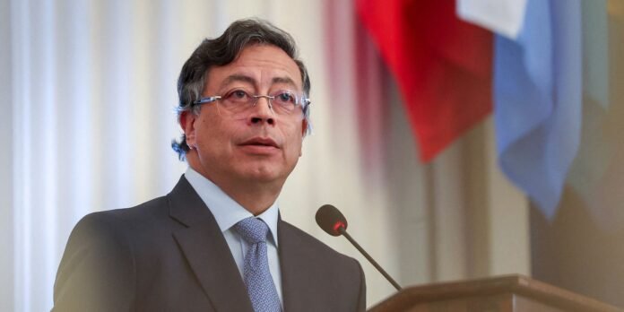 Man in a suit and glasses speaking at a podium with a microphone, with colorful flags in the background.