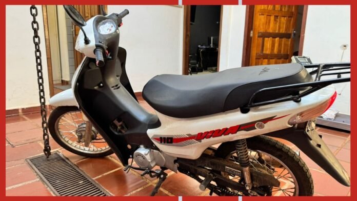 White scooter parked indoors on a tile floor with a red and black stripe design and a rear rack, front chain visible on the left side