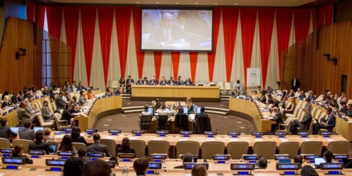 Wide shot of a large international conference room with delegates seated around curved desks and a central panel, a large screen above shows the proceedings, red and white drapery in the background.