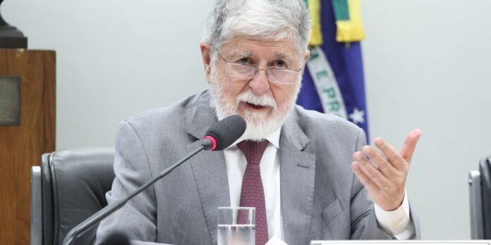 Older man in a gray suit and red tie speaks into a microphone at a formal panel discussion
