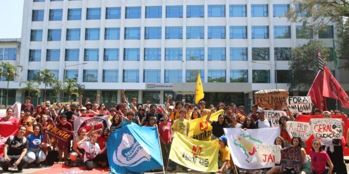 Large group of students holding banners and flags for a campus protest in front of a modern university building.