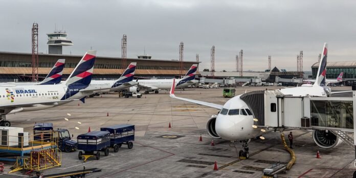 Wide view of an airport apron with several planes at gates; a jet bridge connects to a white airliner in the foreground, with ground equipment and a distant control tower under a cloudy sky.